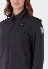 Giacca nera leggera con colletto rialzato, tasche laterali e spalline. Include una patch con il logo sulla manica sinistra.