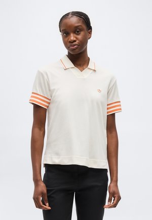 Jauna sieviete ar pītiem matiem, valkā baltu īsroku polo kreklu ar oranžām svītrām uz piedurknēm un apkakles, kā arī melnas bikses.