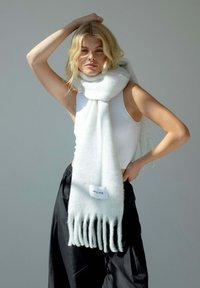 Écharpe en laine blanche avec des bords frangés, ornée d'une étiquette logo portant l'inscription "Arctic Fox". Le mannequin porte un haut sans manches et un pantalon noir ample.