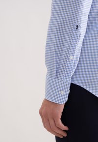 Jasnoniebieska koszula z nadrukiem w kratę, białe guziki, dopasowany mankiet i czarny haft na rękawie. Teksturowana bawełniana tkanina.