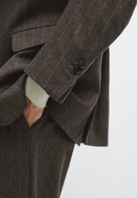 Braun gestreifter Anzug aus strukturiertem Material, mit einem Zweiknopfmanschette, seitlicher Tasche und einem weichen beigen Pullover, der darunter hervorschaut.
