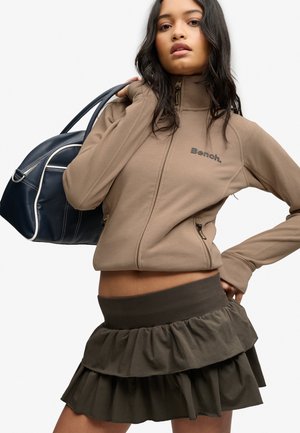 Femme portant une veste marron zippée avec le logo "Bench." et une jupe à volants foncée, tenant un sac à main bleu marine sur l'épaule.
