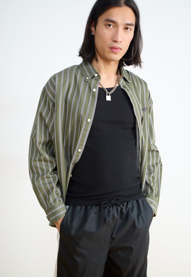 Chemise à rayures vertes boutonnée portée avec un débardeur noir à côtes et un short noir avec cordon de serrage ; collier en chaîne argenté en accent.