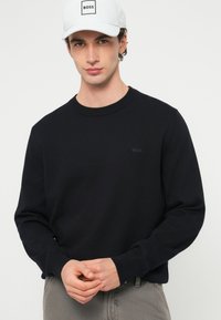 Νεαρός άνδρας που φοράει άσπρο καπέλο με την επιγραφή «BOSS» και μαύρο πουλόβερ με μικρό λογότυπο «BOSS», στέκεται μπροστά σε έναν απλό φωτεινό φόντο.