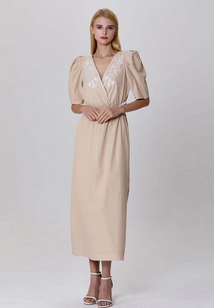 Robe midi beige avec manches bouffantes, ornée de broderies florales blanches le long du décolleté en V. Tissu lisse et taille cintrée. Sandales à bouts ouverts.