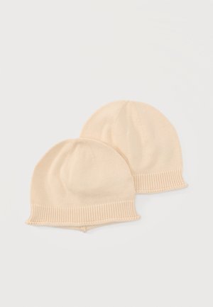 Zwei identische beige gestrickte Babymützen mit gerippten Rändern, die auf einem einfarbigen hellen Hintergrund liegen.