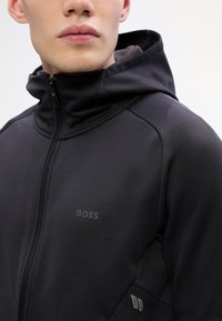 Črna zip-up kapuca z teksturirano tkanino, ki ima kapuco, subtilno blagovno znamko na prsih in edinstveno zasnovo na strani.