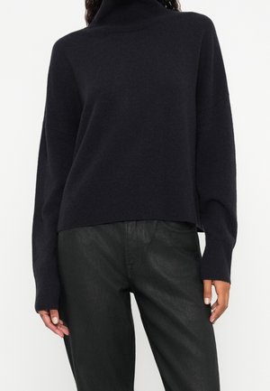 Kobieta ubrana w czarny sweter z golfem z długimi rękawami oraz czarne skórzane spodnie z wysokim stanem, stojąca na tle białego tła.