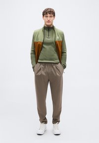 Πράσινο γιλέκο με φερμουάρ και καφέ λεπτομέρειες πάνω από πράσινη μπλούζα μακρυμάνικη, συνδυασμένο με μπεζ παντελόνι και λευκά αθλητικά παπούτσια. Μινιμαλιστικός σχεδιασμός.