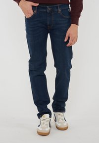 Jeans in denim blu scuro con vestibilità slim, cuciture visibili, tasche anteriori e chiusura con bottone, abbinati a sneakers casual bianche e beige.