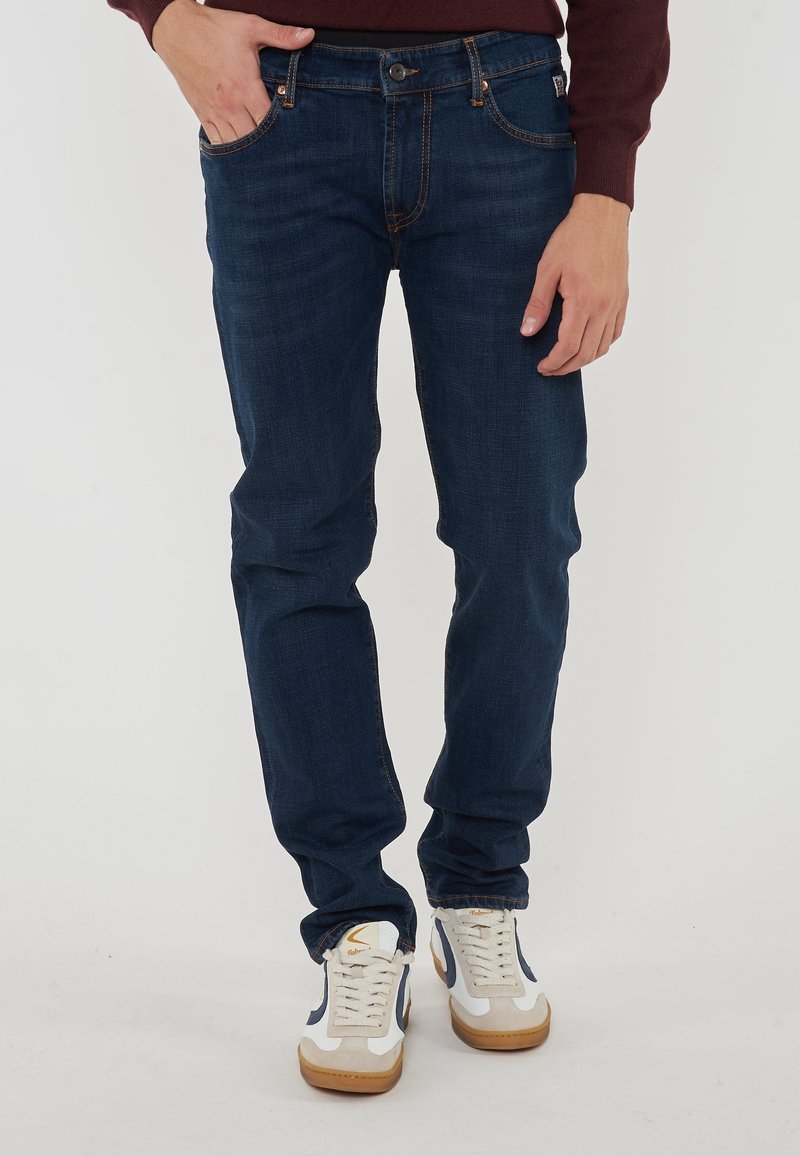 Jeans in denim blu scuro con vestibilità slim, cuciture visibili, tasche anteriori e chiusura con bottone, abbinati a sneakers casual bianche e beige.