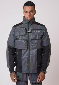 Giacca nera e iridescente con chiusura a zip, colletto alto e molteplici tasche. Presenta un tessuto testurizzato e materiali in contrasto.