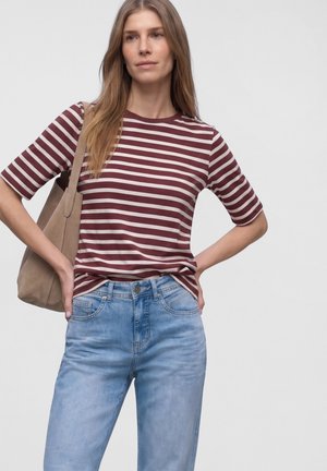 Frau trägt ein bordeauxrotes und weiß gestreiftes T-Shirt, hellblaue Jeans und eine beige Schultertasche, steht mit den Händen in der Hüfte.