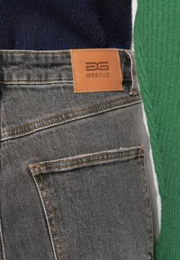 Tmavomodré džínsy z denimu s oranžovým prešitím a hnedou koženou nálepkou s nápisom "GESTUZ." Čiastočne viditeľný zelený žebrovaný sveter.