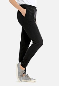 Persoon in zwarte slim-fit broek met de hand in de zak en grijze casual sneakers, staande tegen een effen lichte achtergrond.
