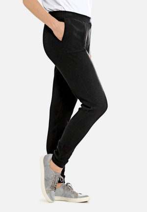 Persoon in zwarte slim-fit broek met de hand in de zak en grijze casual sneakers, staande tegen een effen lichte achtergrond.