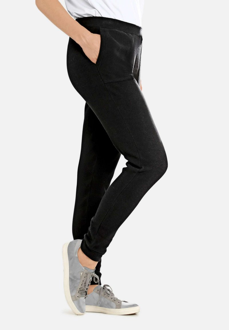 Persoon in zwarte slim-fit broek met de hand in de zak en grijze casual sneakers, staande tegen een effen lichte achtergrond.
