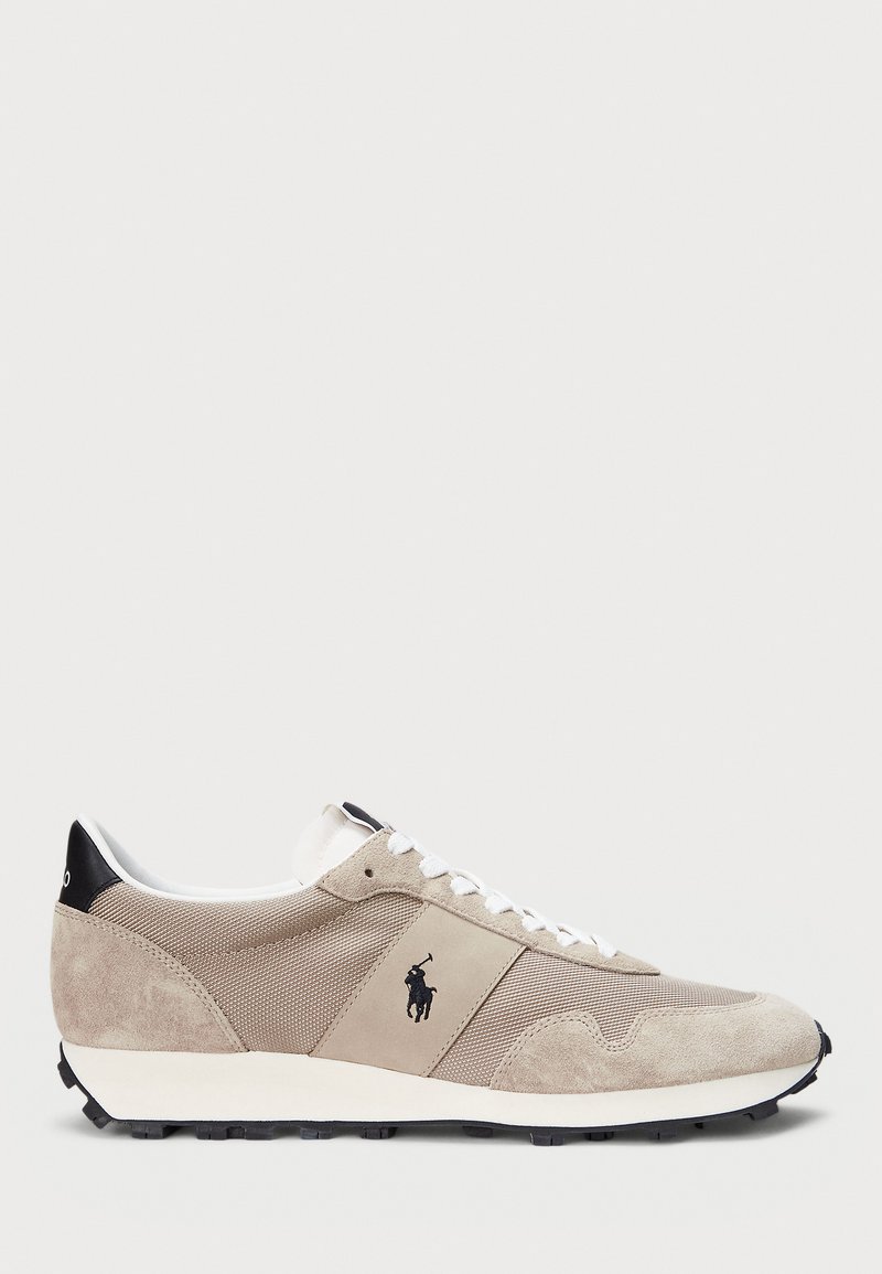 Beige athletic shoe featuring suede and mesh materials, a white sole, black accents, and a logo on the side. Lace-up design with a rounded toe.