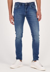 Blå denimjeans med slim fit, subtila slitningar, femficksdesign och en kontrasterande knappstängning. Matchas med vita sneakers.