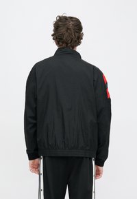 Μαύρο αδιάβροχο μπουφάν με γιακά που στέκεται όρθιος, ελαστική μέση και κόκκινη λεπτομέρεια στον αριστερό ώμο. Ελαφρύ ύφασμα με ματ φινίρισμα.