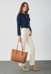 Cardigan en maille bleu marine avec boutons dorés, jean skinny crème, bottes à talons en daim beige et sac fourre-tout marron avec une bordure en fourrure.