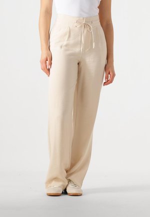 Frau trägt hoch taillierte beige Weitbein-Hose mit gebundenem Gürtel und weißen Turnschuhen, steht vor einem schlichten hellen Hintergrund.