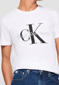 Biała koszulka Calvin Klein Jeans z dużym czarnym logo "cK", noszona z niebieskimi dżinsami, pokazująca górną część torsu oraz część szyi.