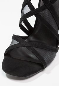 Musta korkeakorkoinen sandaali, jossa on ristikkäiset verkko- ja pehmeän sueden hihnat, sileä pinta ja avoin varvasmuoto.