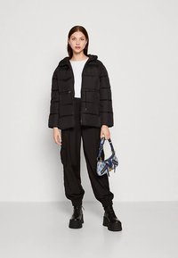 Svart vattert jakke med horisontal quiltet design, glidelåslommer og hette. Brukes over en hvit topp med svarte, vide bukser og platåsko.