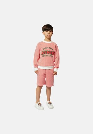 Completo felpa e pantaloni corti a righe rosse e bianche con testo "KENZO", abbinato a sneaker bianche con design di tigre. Texture del tessuto liscia.