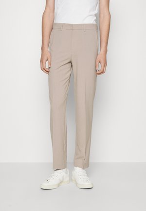 Mężczyzna w beżowych obcisłych spodniach i białych trampkach, stojący na tle jednolitego białego tła.