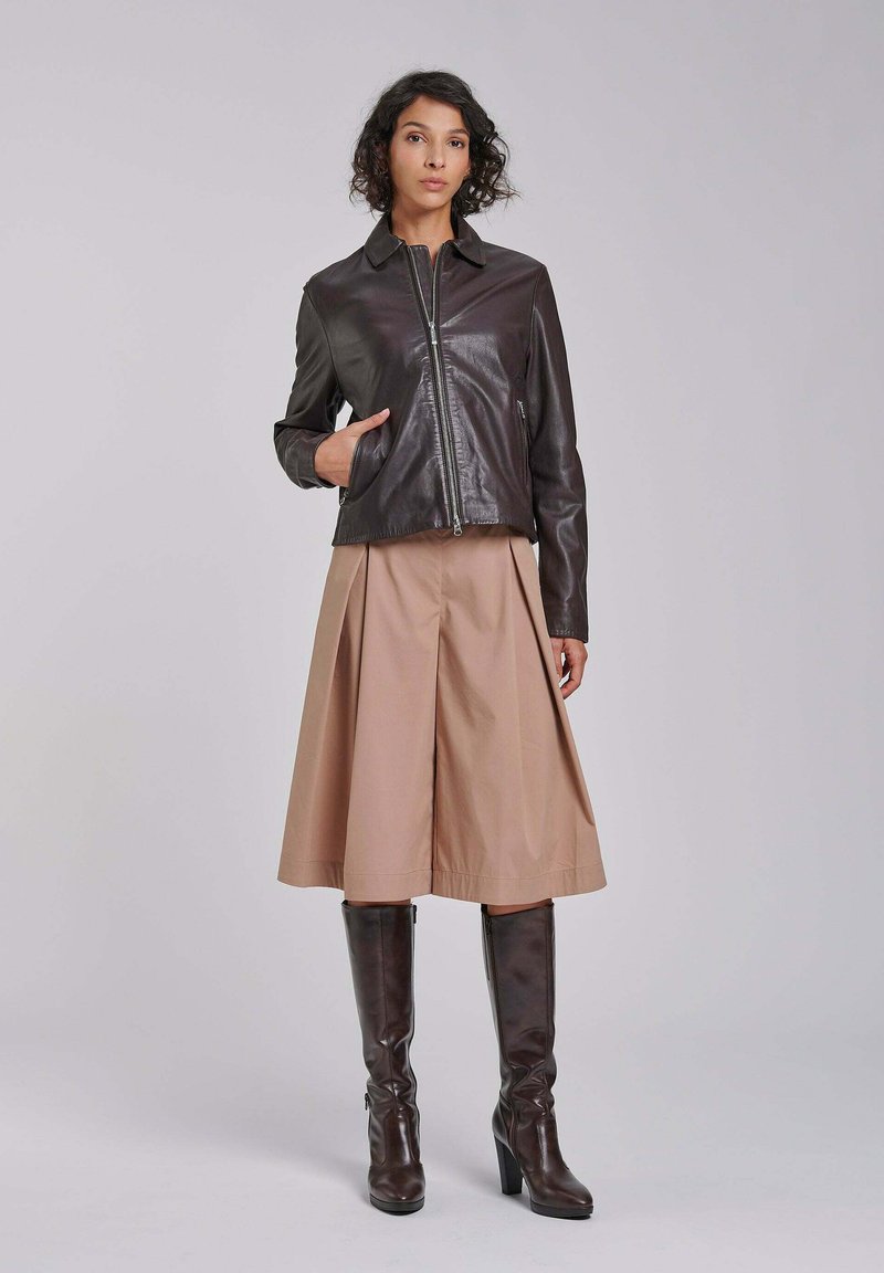 Mujer con chaqueta de cuero marrón oscuro, falda midi beige y botas altas de tacón a juego en marrón oscuro, de pie frente a un fondo liso.