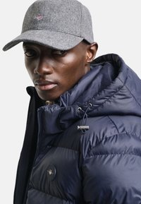 Marineblå puffjakke med quiltet tekstur og justerbar hette, kombinert med en grå ullcap med et lite emblem foran.