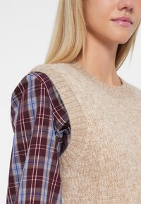 Beige pletený svetr s texturovaným povrchem, doplněný vínově červenou a modrou kostkovanou košilí s nabranými rukávy.