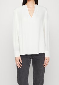Weiße Bluse mit langen Ärmeln, lässiger Passform, V-Ausschnitt und geknöpften Ärmeln. Hergestellt aus einem glatten, leichten Stoff, geeignet für lässige Outfits.