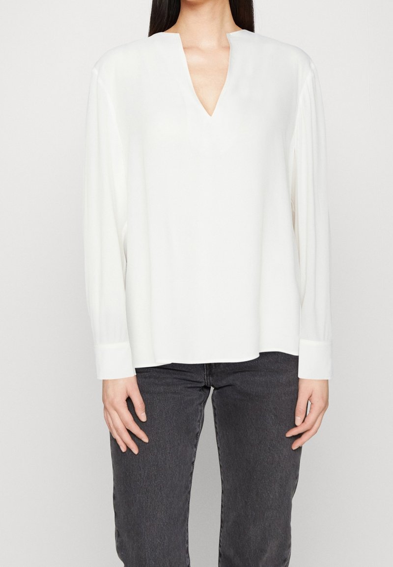 Weiße Bluse mit langen Ärmeln, lässiger Passform, V-Ausschnitt und geknöpften Ärmeln. Hergestellt aus einem glatten, leichten Stoff, geeignet für lässige Outfits.