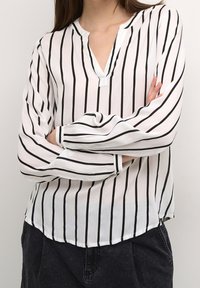 Camicia bianca con righe verticali nere, tessuto leggero, scollatura a V e maniche lunghe con polsini elasticizzati. Dettaglio dell'orlo leggermente curvo.