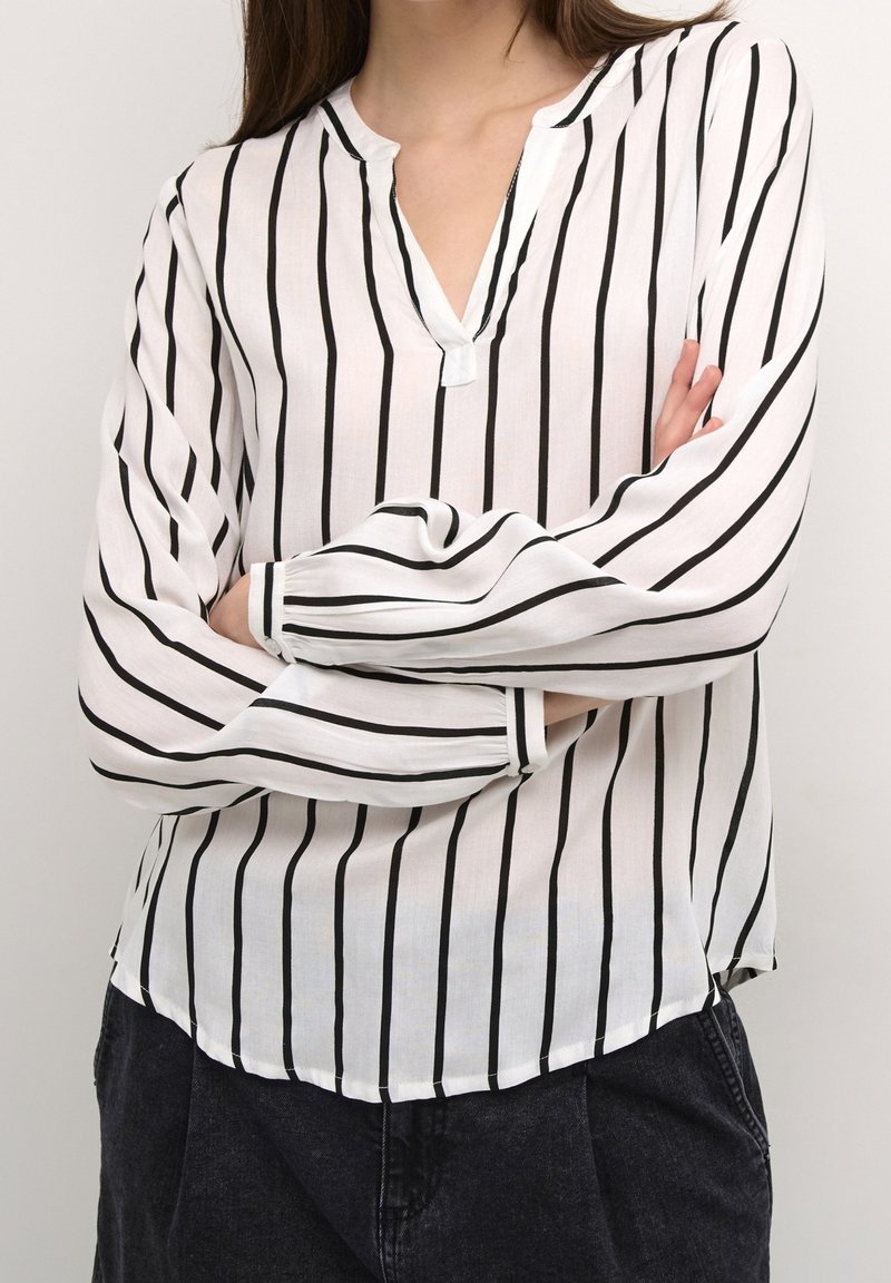 Camicia bianca con righe verticali nere, tessuto leggero, scollatura a V e maniche lunghe con polsini elasticizzati. Dettaglio dell'orlo leggermente curvo.