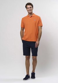 Oranž polo särk kraega ja sinise aktsendilogoga, kombineeritud tumesiniste lühikestega. Modell kannab tumesiniseid tossusid. Ühtlane taust.