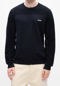 Homme portant un pull noir à manches longues avec des épaules texturées et le logo "BOSS" sur la poitrine, associé à un pantalon beige.