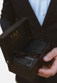 Schwarze Geschenkbox mit goldenem Logo, die eine gemusterte Geldbörse, einen silbernen Schlüsselanhänger und ein Band enthält. Die Box hat eine glatte Außenseite und einen klappbaren Deckel.