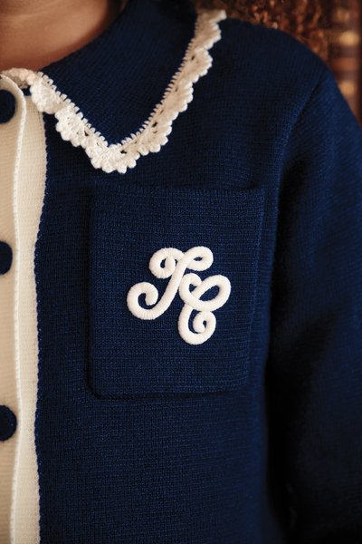 Cardigan azul-marinho com acabamento em colarinho branco, fechamento por botões e um pequeno bolso com iniciais bordadas em branco. Tecido de textura suave.