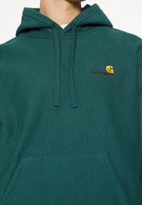 Mørk teal hættetrøje lavet af en bomuldsblandingsstof, som har en front kangaroo lomme, justerbar snor i hætten og broderet logo i gul og rød.