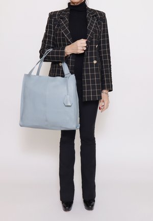 Sac fourre-tout en cuir bleu clair à texture lisse, avec une étiquette hexagonale et deux poignées. Porté avec un blazer et un pantalon à carreaux noirs.