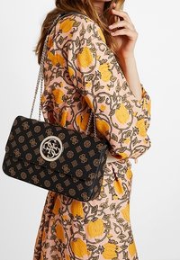 Sac à main noir texturé avec un fermoir monogrammé doré, une bandoulière chaîne et une forme structurée ; présenté devant une robe à motif floral.