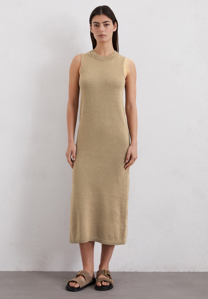 Robe en tricot sans manches, beige, avec un col et un ourlet côtelés. Le tissu a une texture douce. Associée à des sandales brunes à bout ouvert.