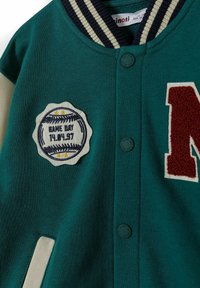 Grön tyg varseljacka med en ribbad krage i marinblått och krämfärgat, en strukturerad röd "N" och en rund broderad emblem med baseballdesign.