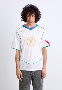 Camiseta de fútbol blanca con un diseño a cuadros, cuello azul y rojo, acentos verdes a lo largo de las costuras, y un logo circular dorado con el número "90".