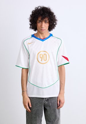 Vit fotbollströja med rutmönster, blå och röd krage, gröna detaljer längs sömmarna och en gul cirkulär logotyp med "90."