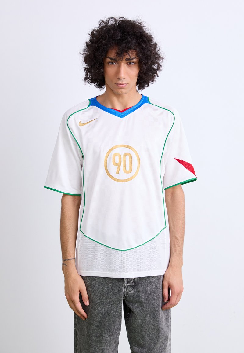 Camiseta de fútbol blanca con un diseño a cuadros, cuello azul y rojo, acentos verdes a lo largo de las costuras, y un logo circular dorado con el número "90".